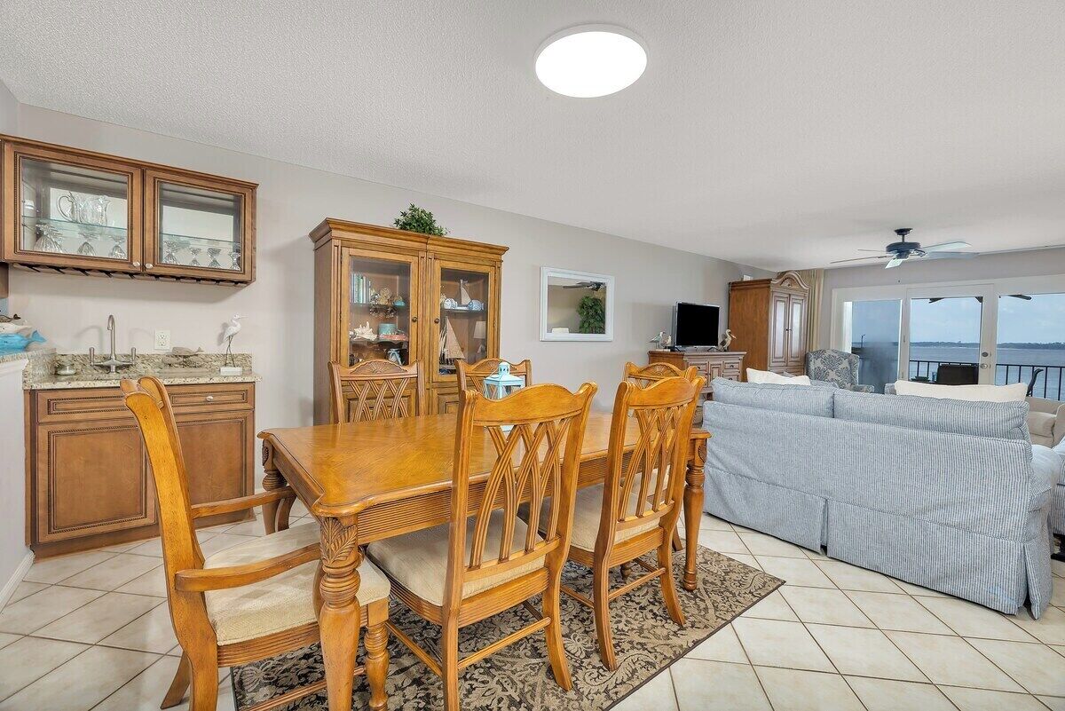 Un coin repas avec une table et des chaises en bois, une cuisine équipée de placards et d’un évier, un salon avec un canapé, et une vue sur la mer grâce à de grandes fenêtres.