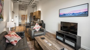 Een moderne woonkamer met een bank, salontafel en televisie. Achtergrond toont een keuken met houten kasten en een eethoek.