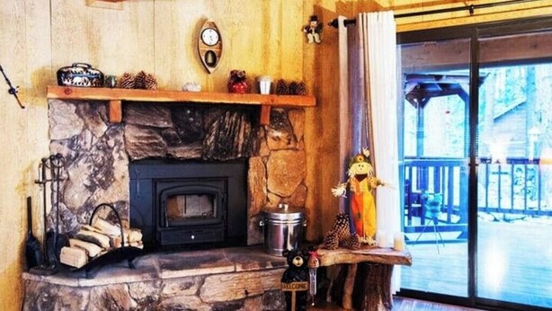 A cozy cabin interior with a stone fireplace, wooden shelves, and a large window overlooking a deck.