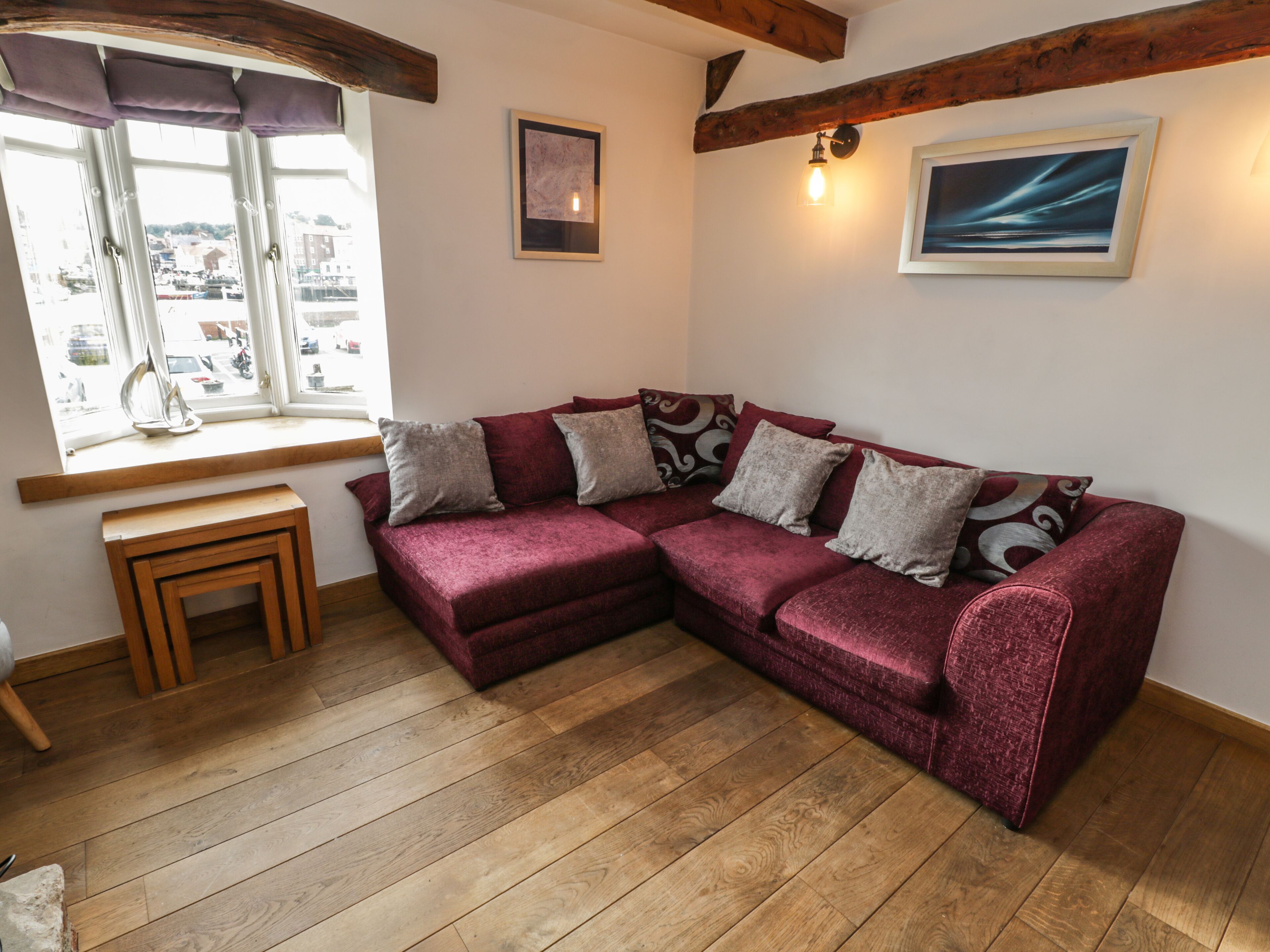 Una stanza accogliente con pavimento in legno, un divano componibile rosso con cuscini decorati, una piccola panca in legno e una finestra con vista sugli edifici.