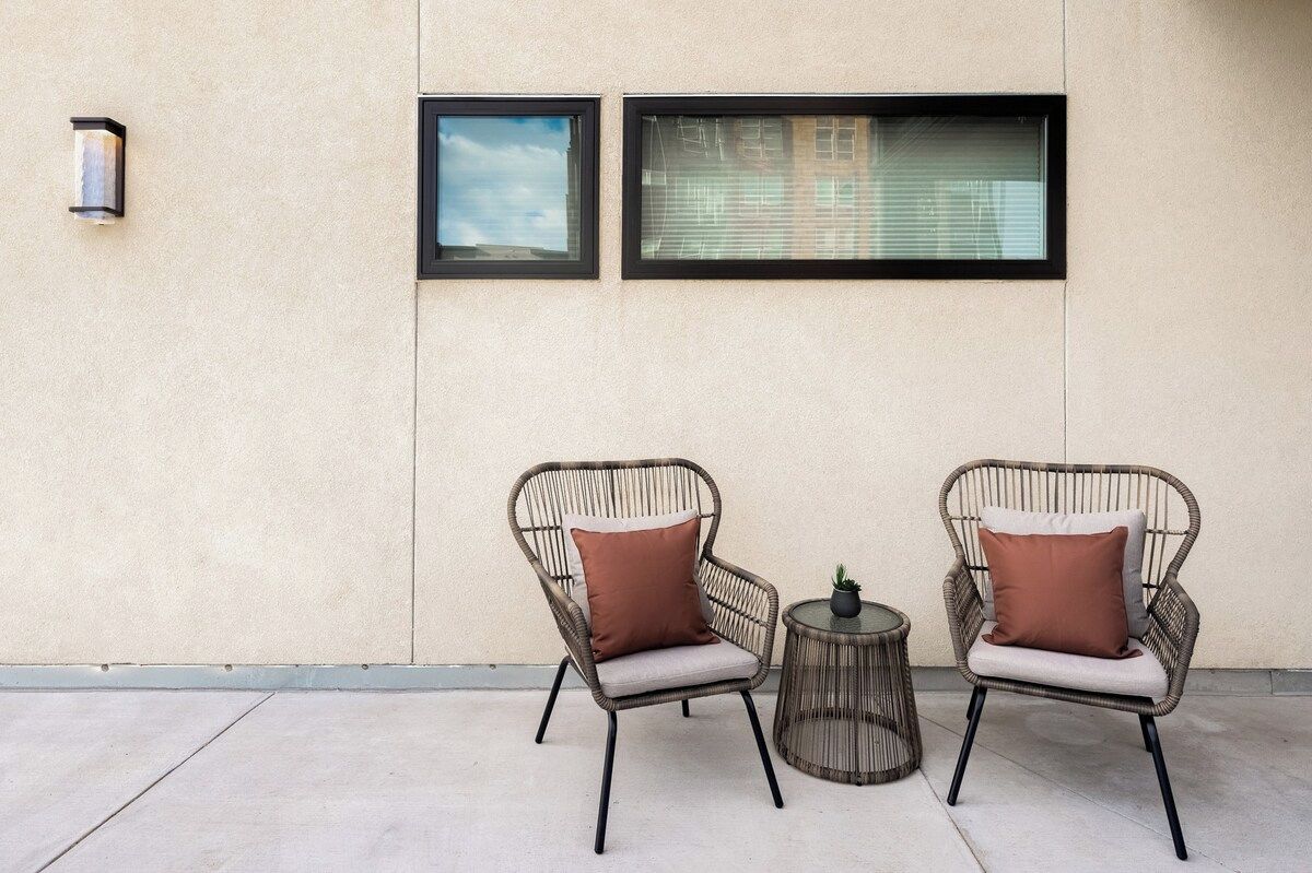 Two wicker chairs with cushions, a small round table, and a potted plant, positioned against a wall with two windows.