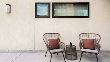 Two wicker chairs with cushions, a small round table, and a potted plant, positioned against a wall with two windows.