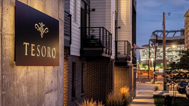 Een bord met de naam 'TESORO' hotel, met een modern ontwerp, op een straat met een gebouw van baksteen en een glazen constructie op de achtergrond.