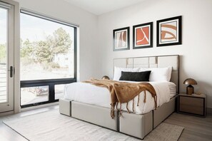 Un dormitorio con cama, mesita de noche, lámpara y ventana que ofrece vistas a árboles y un edificio.