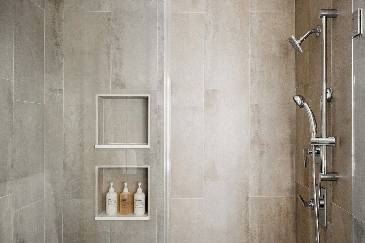 Un baño con ducha, dos repisas blancas con artículos de aseo y una pared con azulejos.