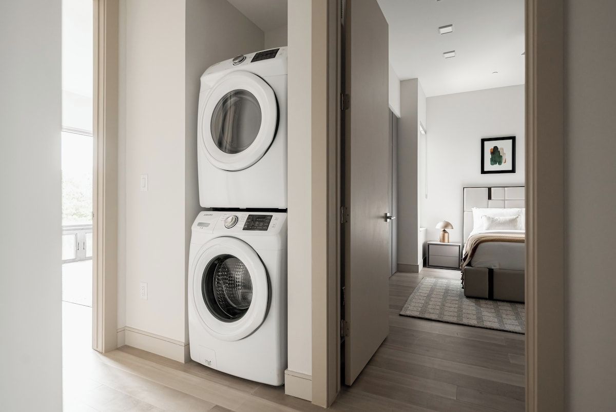 A laundry room with two stacked washing machines and a bedroom with a bed and nightstand.