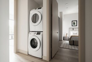 A laundry room with two stacked washing machines and a bedroom with a bed and nightstand.