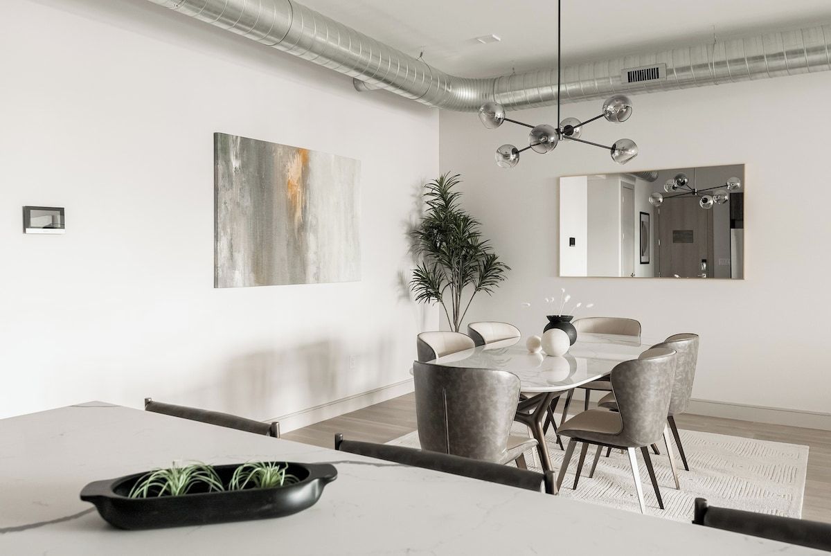 Un espace de salle à manger moderne avec une table ronde, des chaises recouvertes d’un tissu gris, une plante en pot et un lustre contemporain.