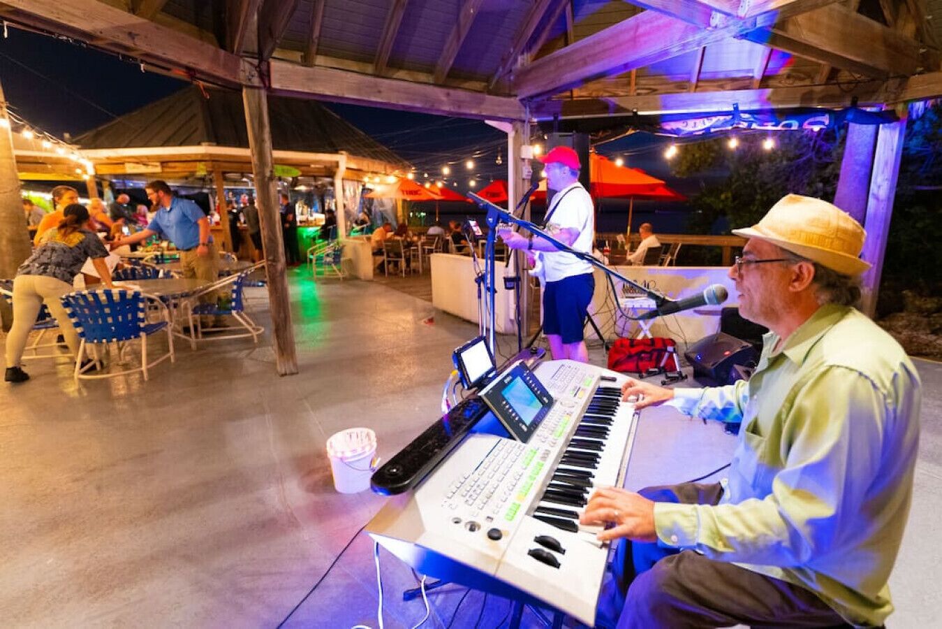 Une personne joue du clavier lors d’un événement en plein air, où sont disposées des tables et des chaises, sous une zone abritée et illuminée par des guirlandes lumineuses.