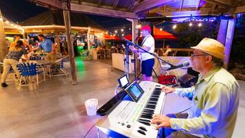 Une personne joue du clavier lors d’un événement en plein air, où sont disposées des tables et des chaises, sous une zone abritée et illuminée par des guirlandes lumineuses.