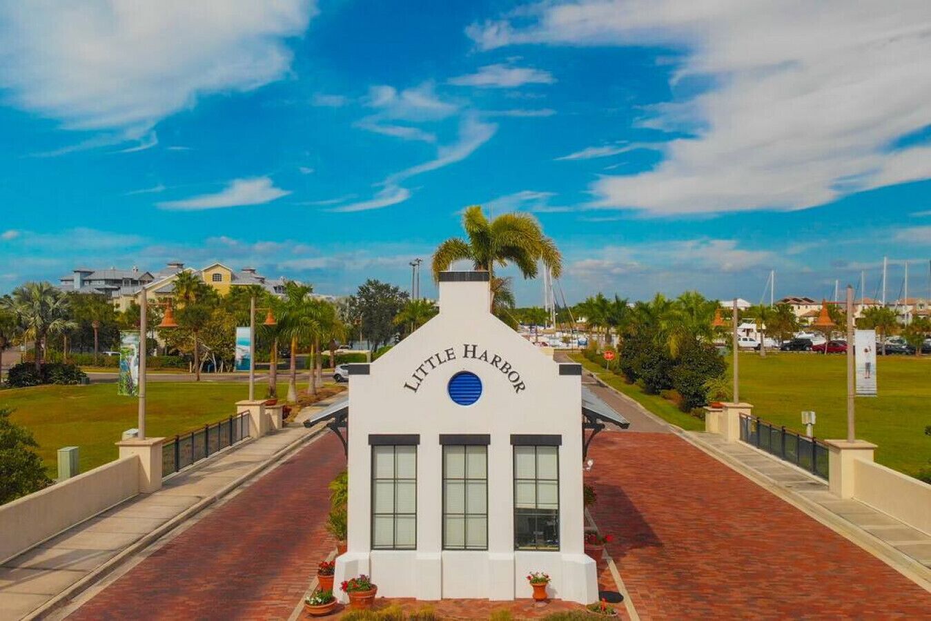 A white building with the sign "Little Harbor" on a brick pathway with palm trees and a clear blue sky in the background.