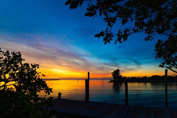 Vista al atardecer desde un muelle de madera, con agua tranquila y tierra a lo lejos.