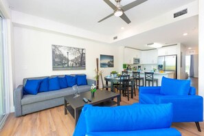 Una sala de estar moderna con un sofá gris y sillones azules, un comedor con mesa y sillas, y una cocina al fondo.