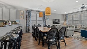 Eine moderne Küche mit weißen Schränken, eine Essecke mit Holztisch und schwarzen Stühlen sowie ein Wohnzimmer mit grauem Sofa und gemusterten Vorhängen.