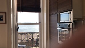 A room with a view of the sea through a window with a metal railing. There are built-in appliances, including a microwave and oven, on the wall. - Apartment - Sleeps 4 - Balcony - Sea View (Eastbourne)