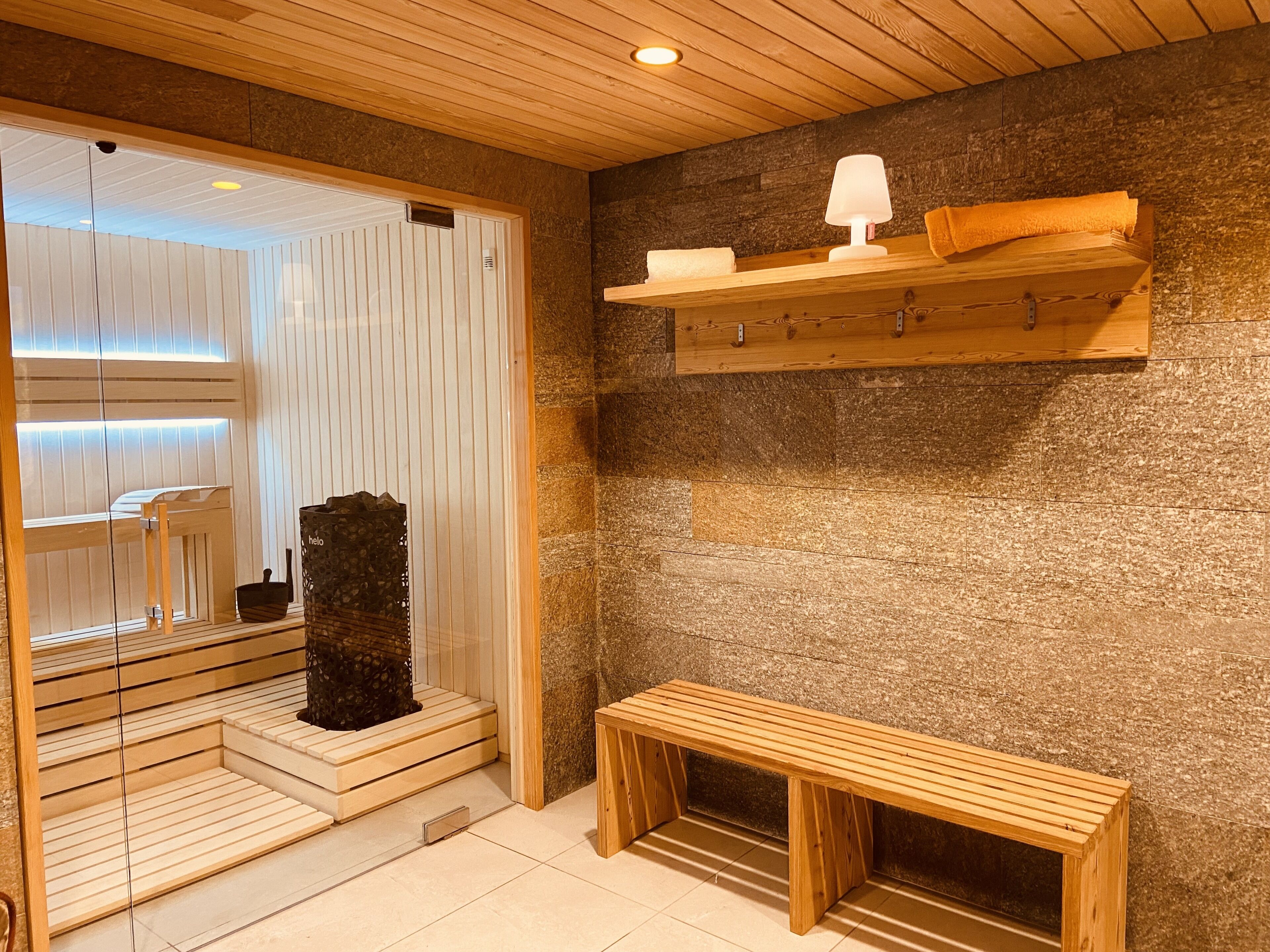 A sauna room with wooden benches, a bench with a towel, a heater, and a shelf with a towel and a lamp.