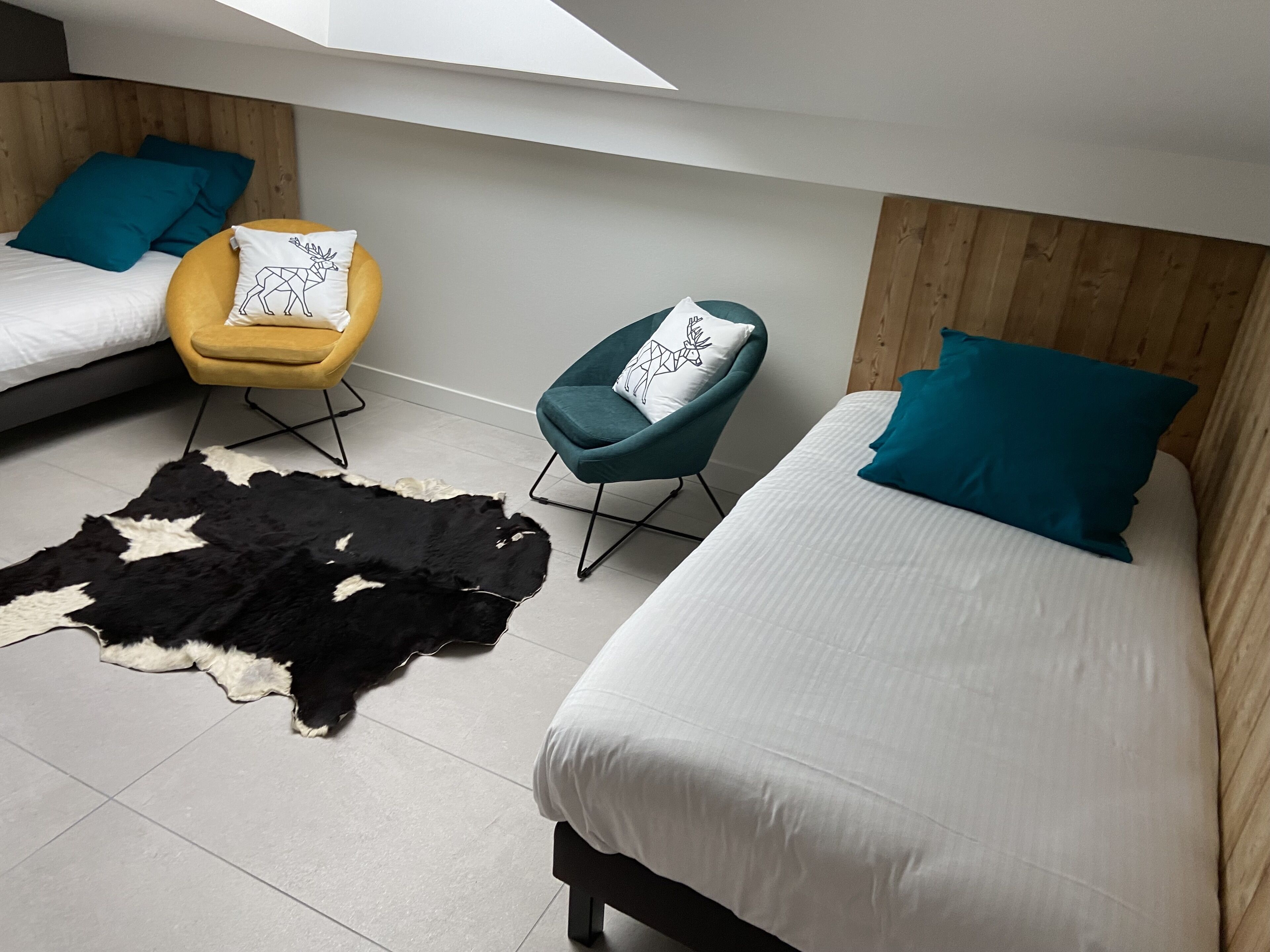 Un dormitorio moderno con dos camas, una silla amarilla, una silla color turquesa y una alfombra de cuero de vaca.