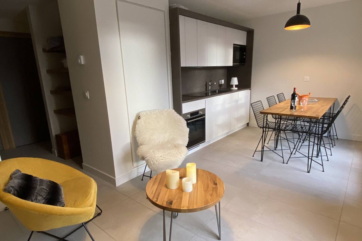 Una sala de estar moderna con un sillón amarillo, una mesa redonda de madera con velas, un sofá azul y un comedor con mesa y sillas de madera.