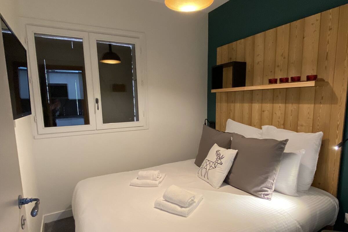 Una cama bien hecha con sábanas blancas y un cojín decorativo, una mesita de noche, una lámpara colgante y una ventana con vistas.