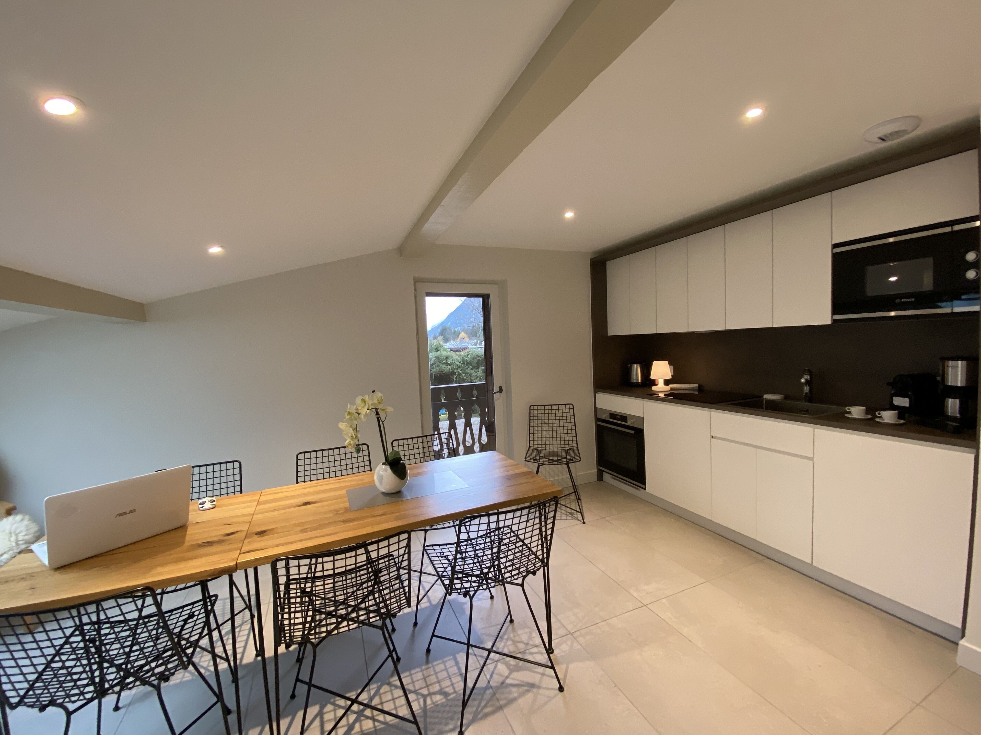A modern kitchen with white cabinets, a wooden dining table, and a laptop on the table.