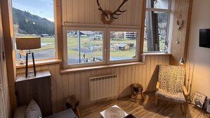 Ein gemütliches Zimmer mit Holzboden, einem großen Fenster mit Blick auf einen Wald, einem aufgehängten Hirschgeweih an der Wand, einem Sofa, einem Sessel und einem kleinen Tisch mit einer Lampe.