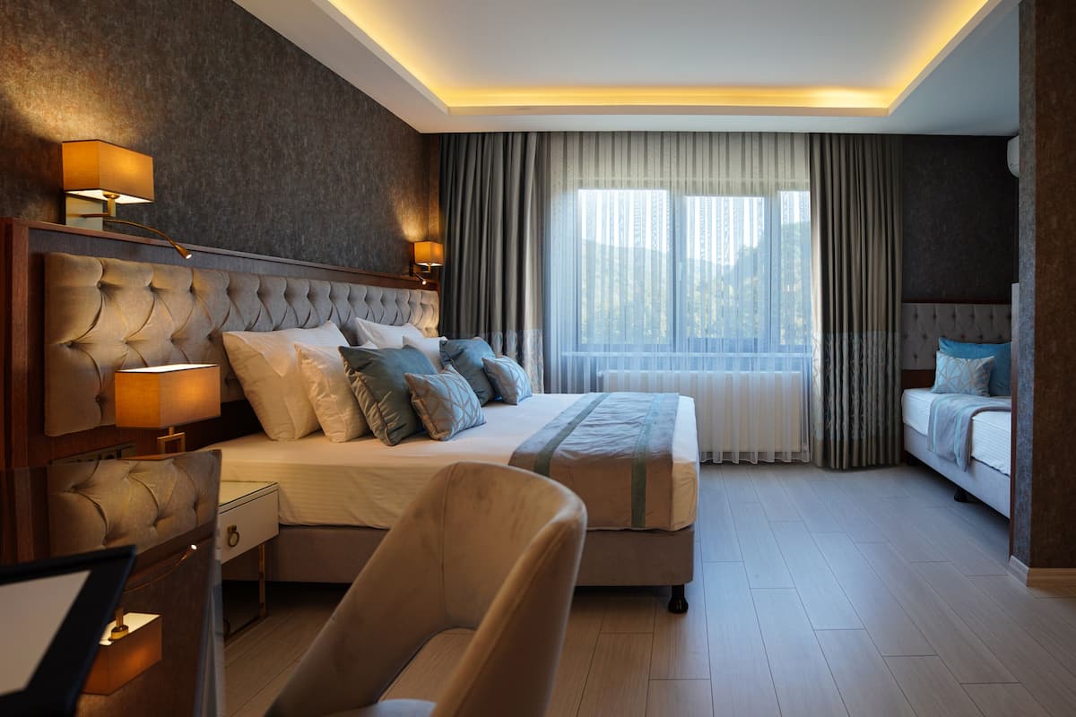 Ett modernt hotellrum med en stor säng, två sängbord med lampor, en stol, ett skrivbord och ett stort fönster med gardiner.