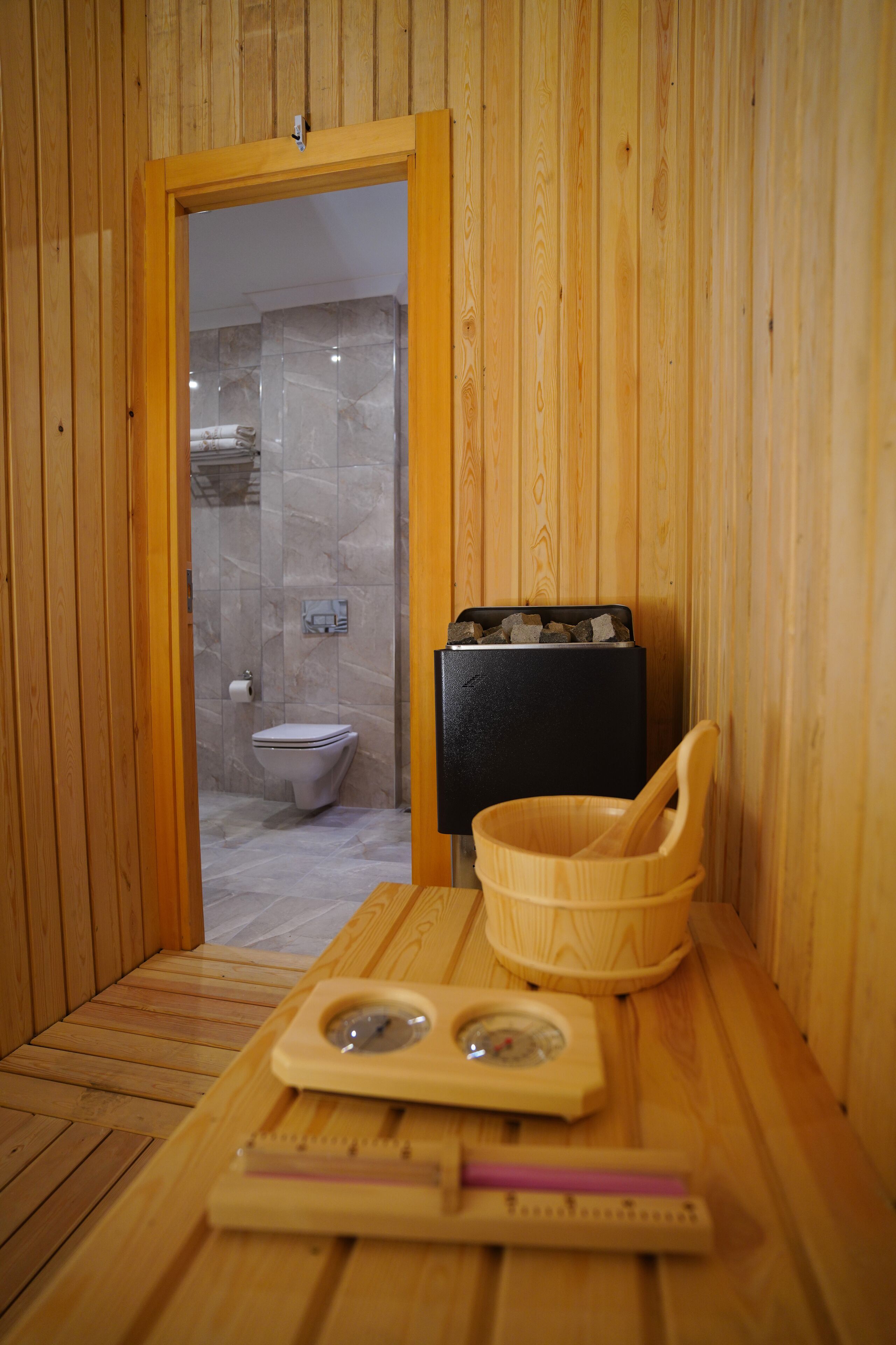 A wooden sauna room with a wooden table, a wooden bowl, a black heater, and a toilet visible through a glass door.