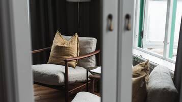 Una habitación con piso de madera, un sillón gris con un cojín color beige, una pequeña mesa y una ventana que ofrece vistas al exterior.