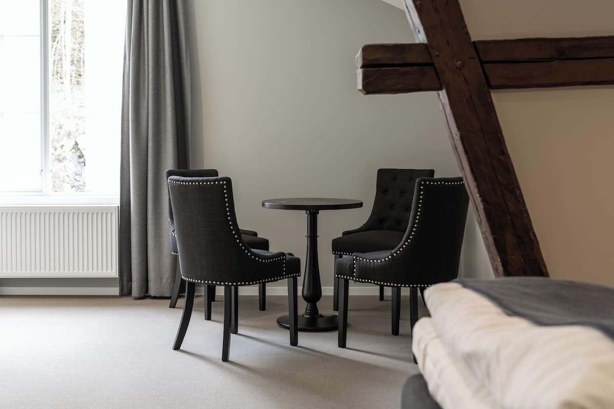A room with a bed, a table, and two chairs. There is a window with curtains, a radiator, and a wooden beam.