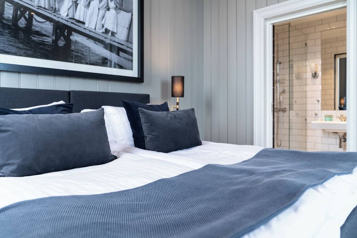 A neatly made bed with dark and light pillows, a framed picture on the wall, and a bathroom visible through an open door.