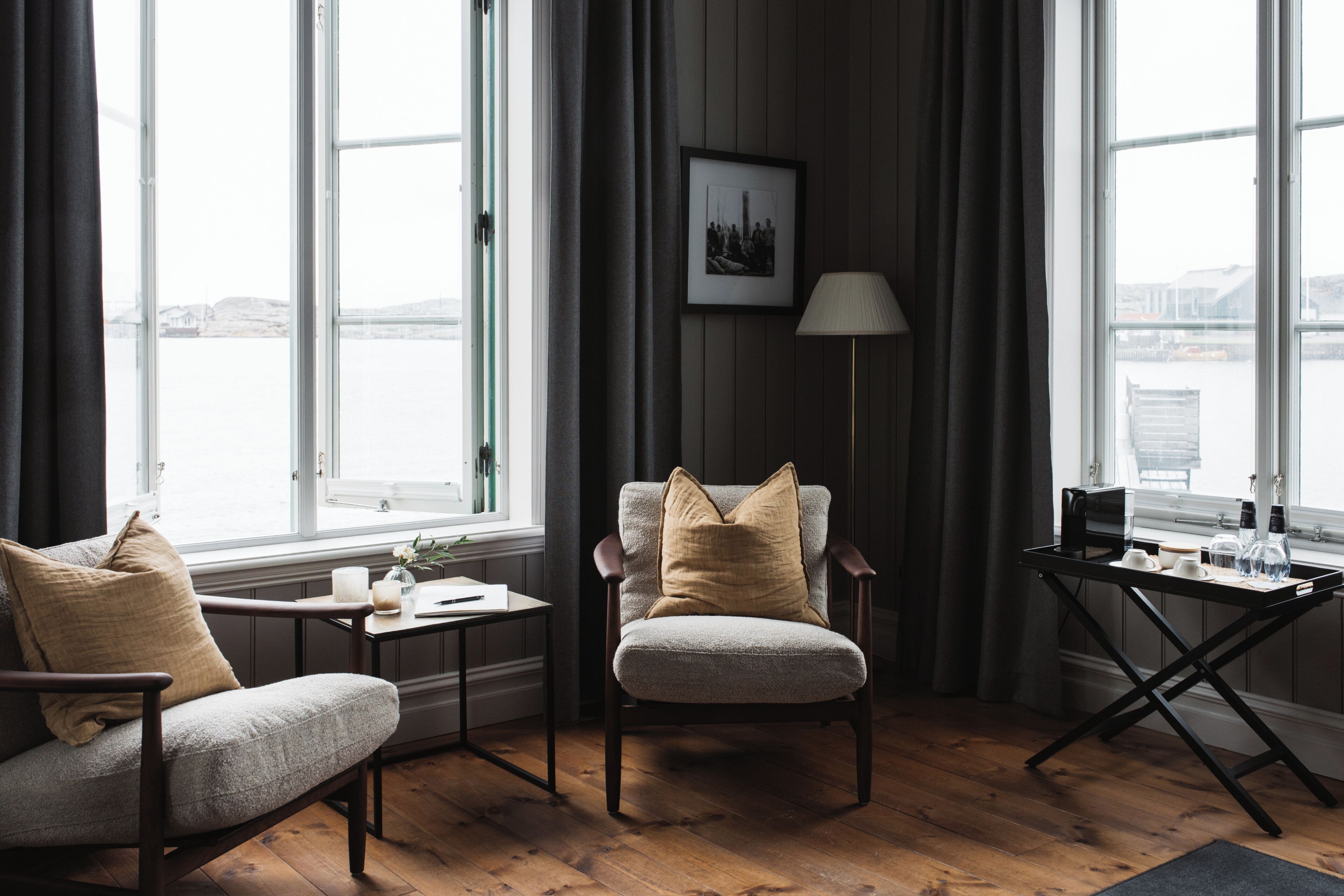 A room with a sofa, armchair, small table, lamp, and a view of the water.