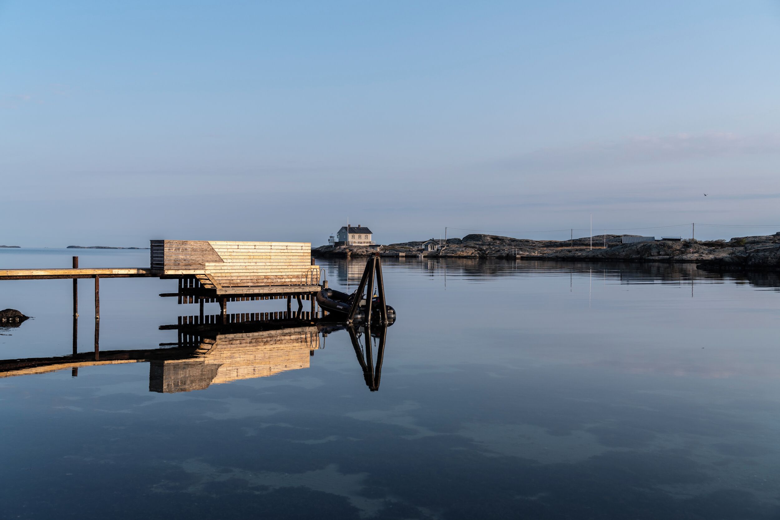 Una orilla tranquila con un muelle de madera que se adentra en el agua, un pequeño edificio a lo lejos y un cielo despejado.