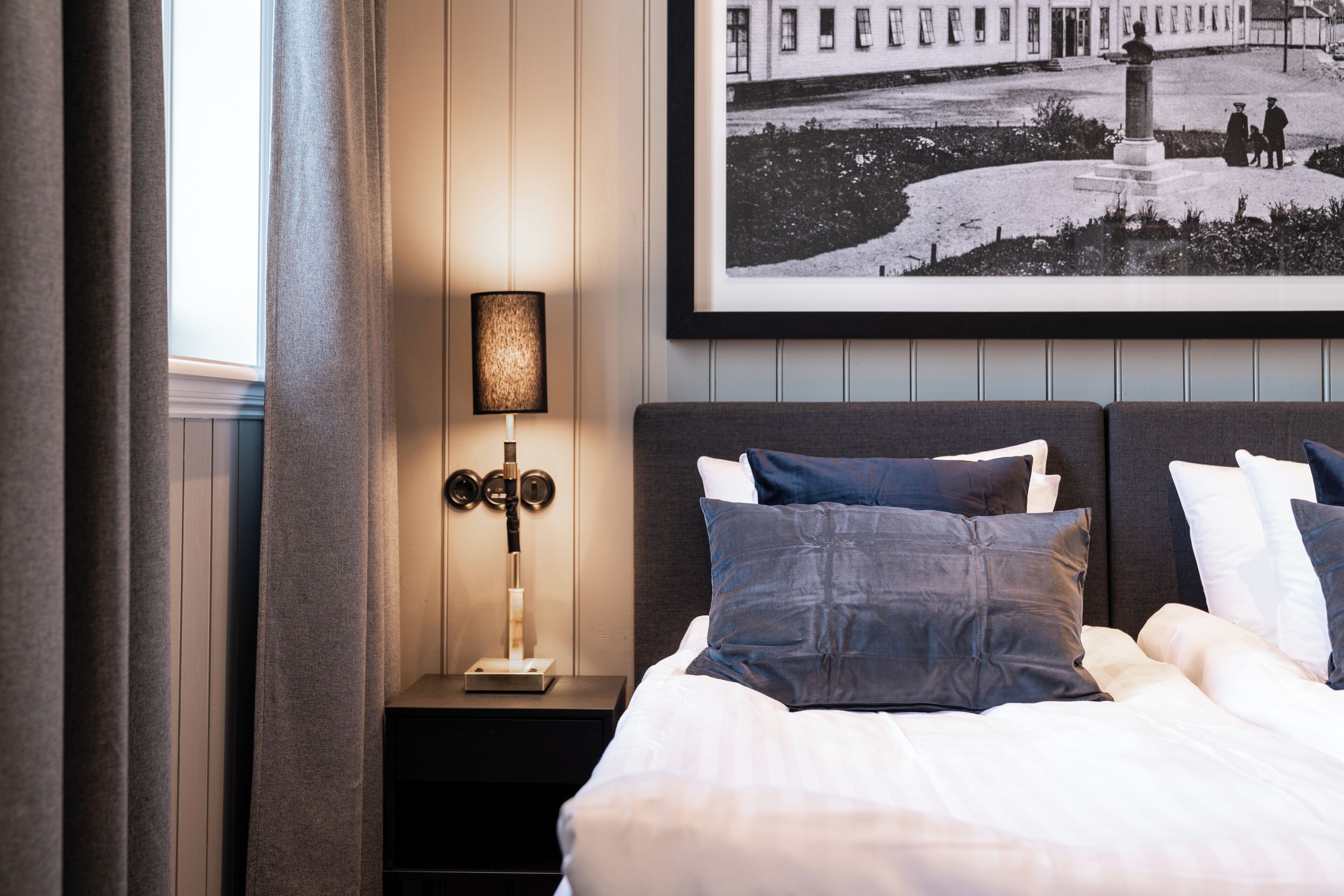 Una cama bien hecha con almohadas oscuras y claras, una lámpara de noche y una fotografía en blanco y negro enmarcada en la pared.