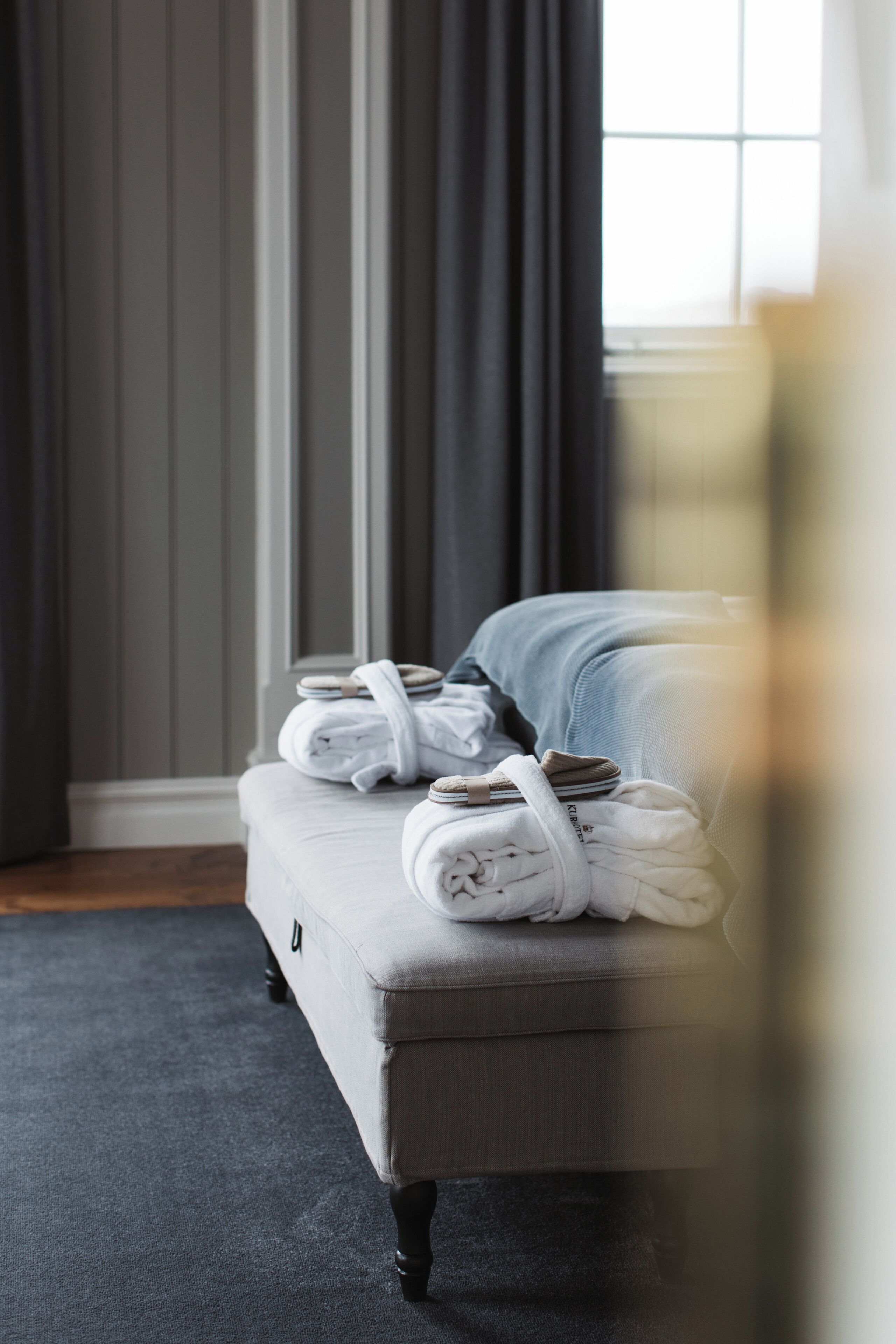 A neatly made bed with a grey headboard, a window with curtains, and a dark floor.