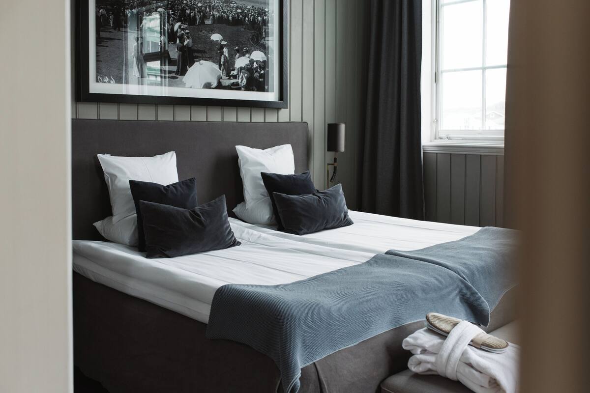 A neatly made bed with white linens and dark pillows, a grey blanket, and a rolled towel on a bedside table.