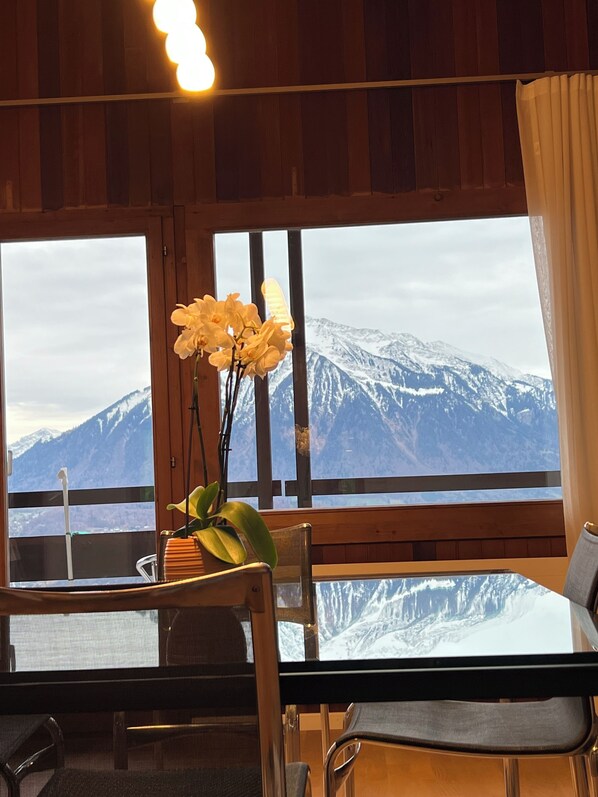 A room with a wooden interior, a table with chairs, a vase with flowers, and a large window offering a view of snow-capped mountains.