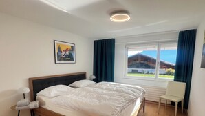 Un dormitorio con cama, una silla, un escritorio, una ventana con cortinas, un cuadro en la pared y una lámpara en el techo.