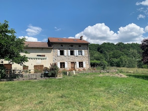 A two-story stone house with a tiled roof, surrounded by a grassy field and a forested hill. - Back in the past (roche en regnier)