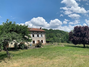 A stone building with a red-tiled roof, surrounded by a grassy field and trees.
