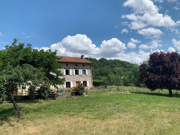 A stone building with a red-tiled roof, surrounded by a grassy field and trees. - Back in the past (roche en regnier)