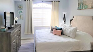 Une chambre à coucher avec un lit, une commode, un téléviseur et une fenêtre avec des rideaux.