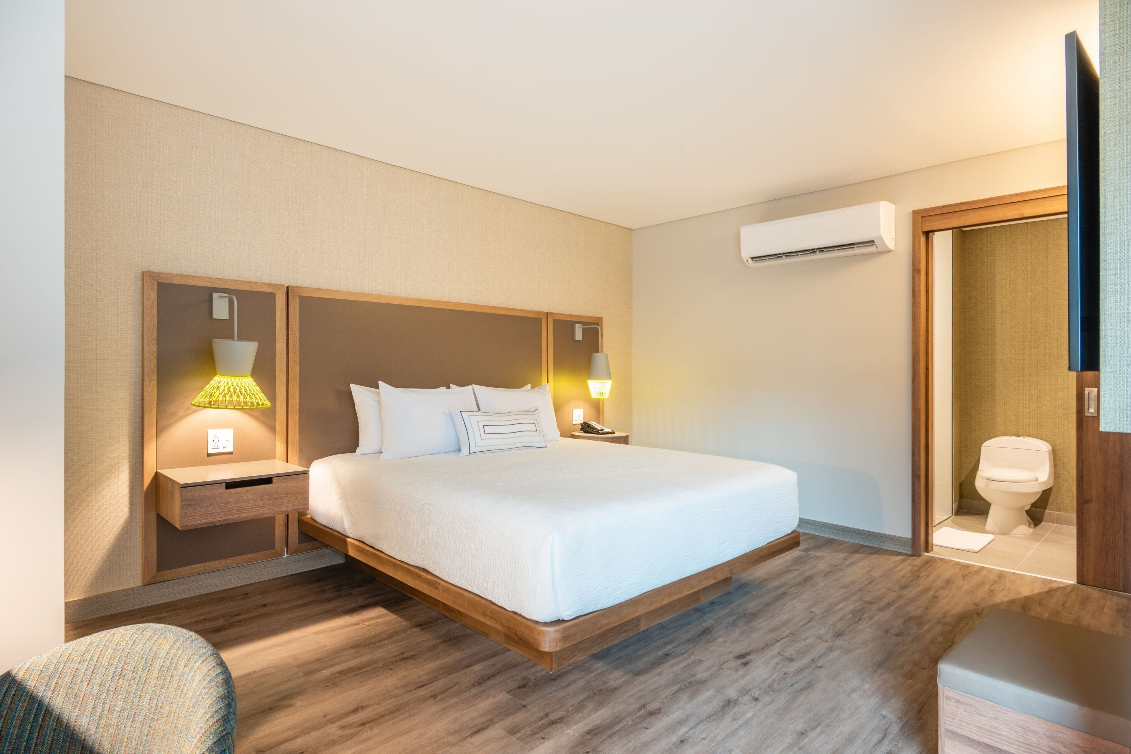 A hotel room with a large bed, wooden headboard, bedside tables with lamps, an air conditioning unit, and a bathroom with a toilet and sink.