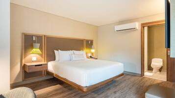 A hotel room with a large bed, wooden headboard, bedside tables with lamps, an air conditioning unit, and a bathroom with a toilet and sink.