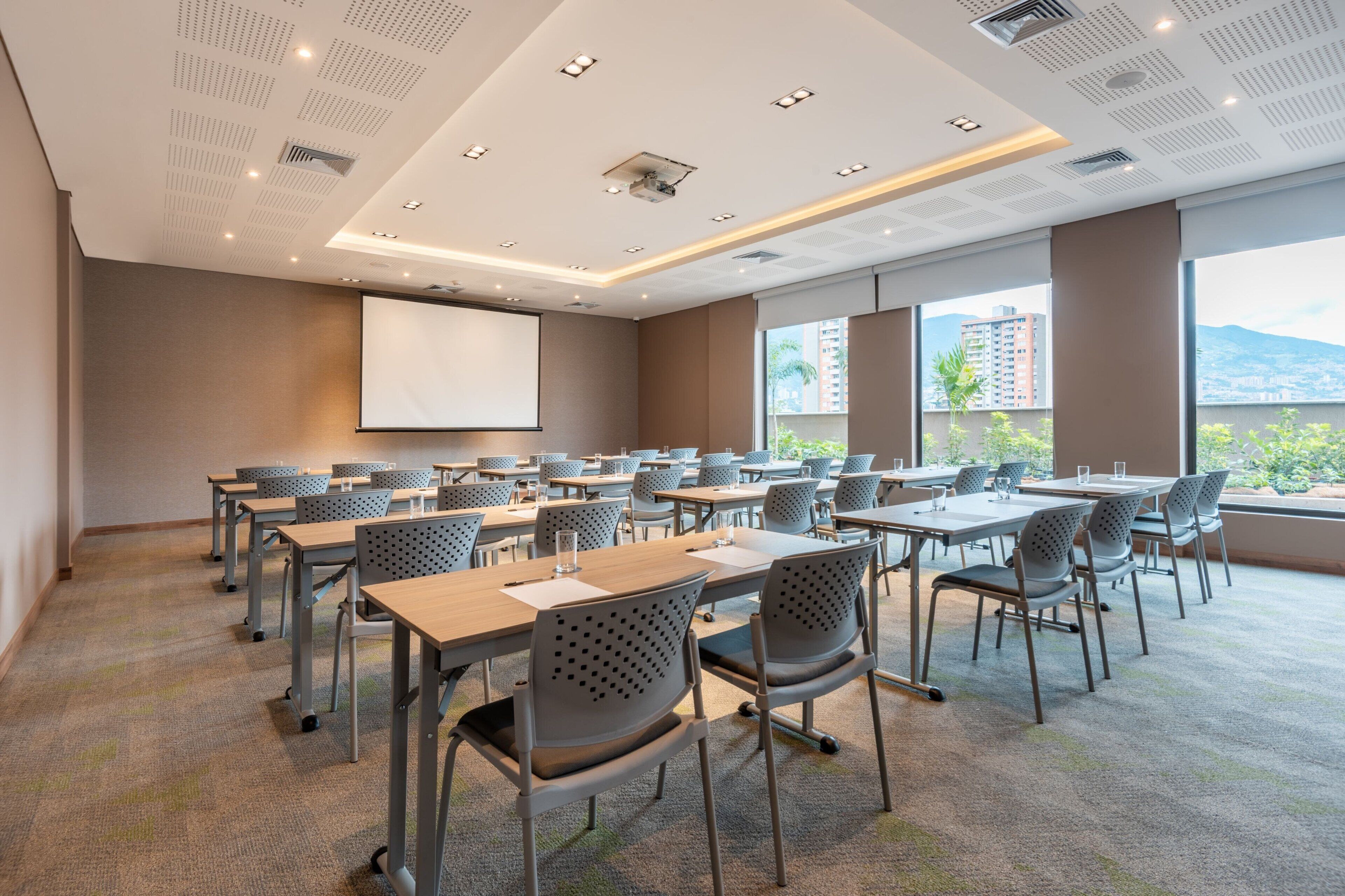 A conference room with multiple tables and chairs, a projector screen, and large windows with a city view.