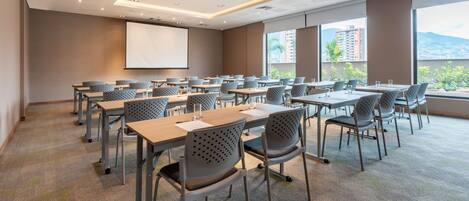 A conference room with multiple tables and chairs, a projector screen, and large windows with a city view.
