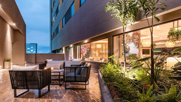 An outdoor patio area with wicker furniture, a brick floor, and a view of a building with large windows and indoor lighting. - Fairfield by Marriott Medellin Sabaneta (Sabaneta)