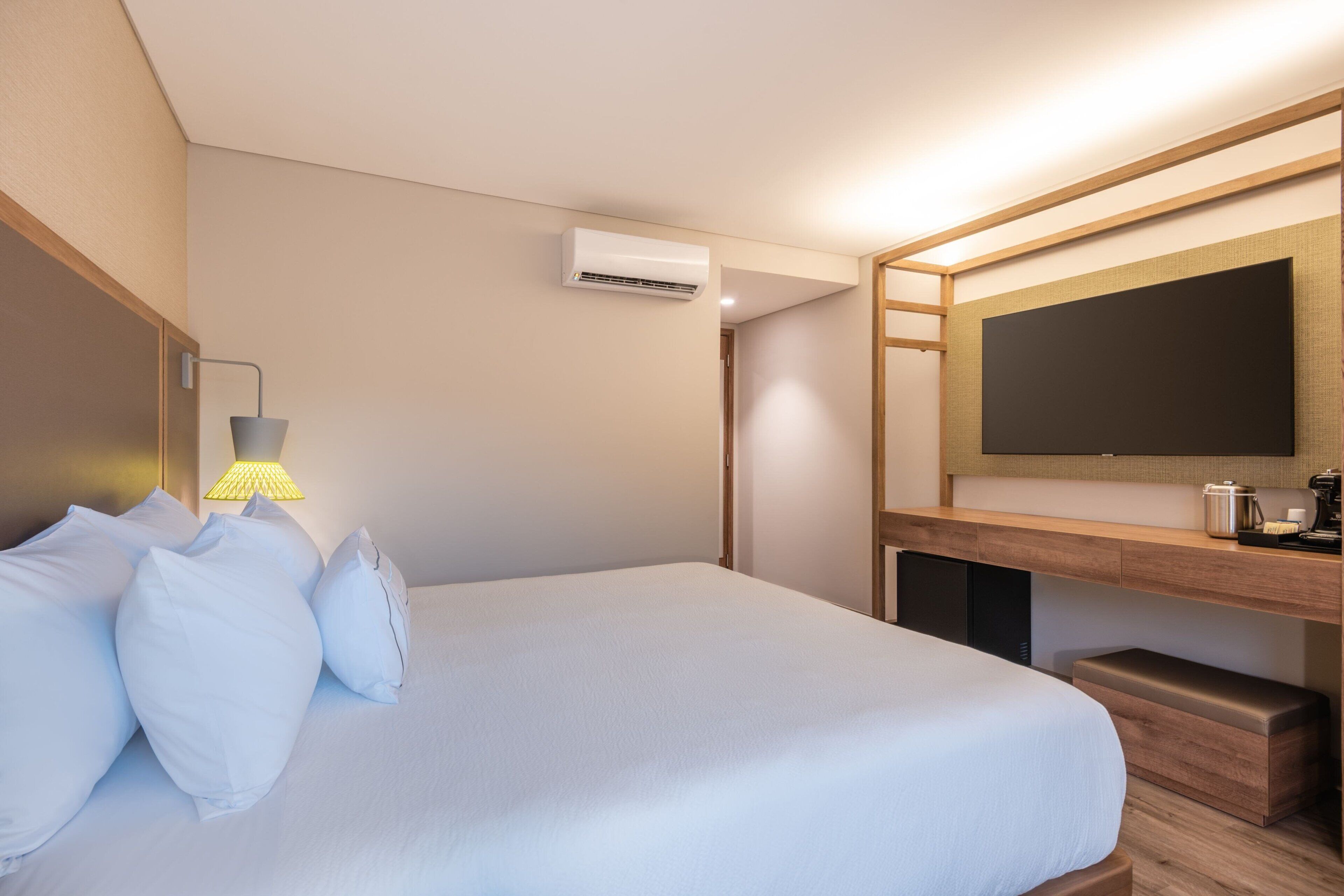 A hotel room with a bed, a bedside lamp, an air conditioning unit, a wooden desk, and a television.