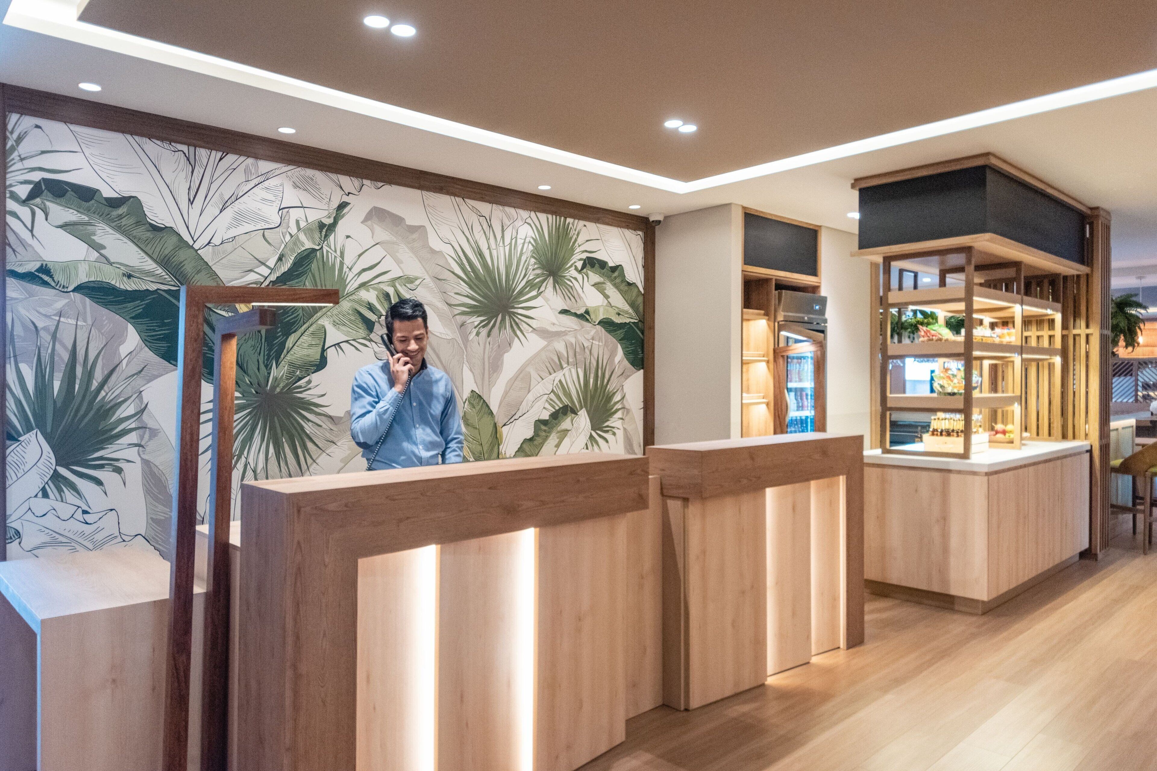 A reception area with a wooden reception desk, a wall with a tropical leaf mural, and a shelving unit with various items.