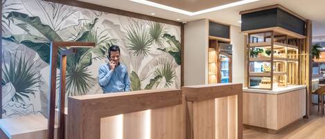 A reception area with a wooden reception desk, a wall with a tropical leaf mural, and a shelving unit with various items.