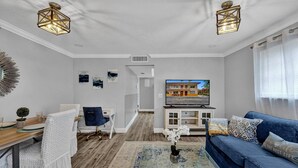 Ein modernes Wohnzimmer mit einem blauen Sofa, einem weißen Sessel, einem Holztisch und einem Fernseher auf einem weißen Schrank. Zwei Pendelleuchten, eine kleine Pflanze und Kunstwerke schmücken die Wände.
